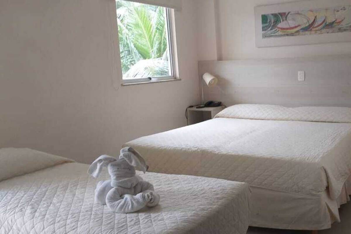 A hotel room with two beds, a window with a view of greenery, a bedside table with a lamp, and a colorful abstract painting on the wall.