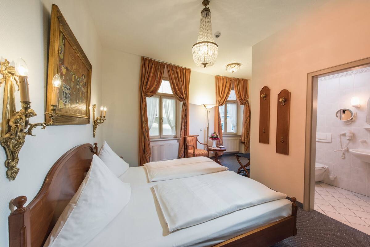 Habitación de hotel con una cama grande, un escritorio, una silla, una araña y un baño visible a través de una puerta abierta.