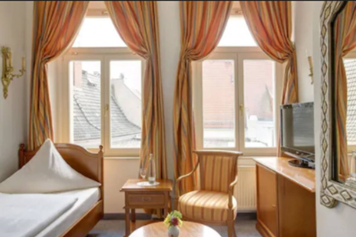 Una habitación de hotel con cama, una mesita con un florero, una silla, un televisor y ventanas con cortinas.