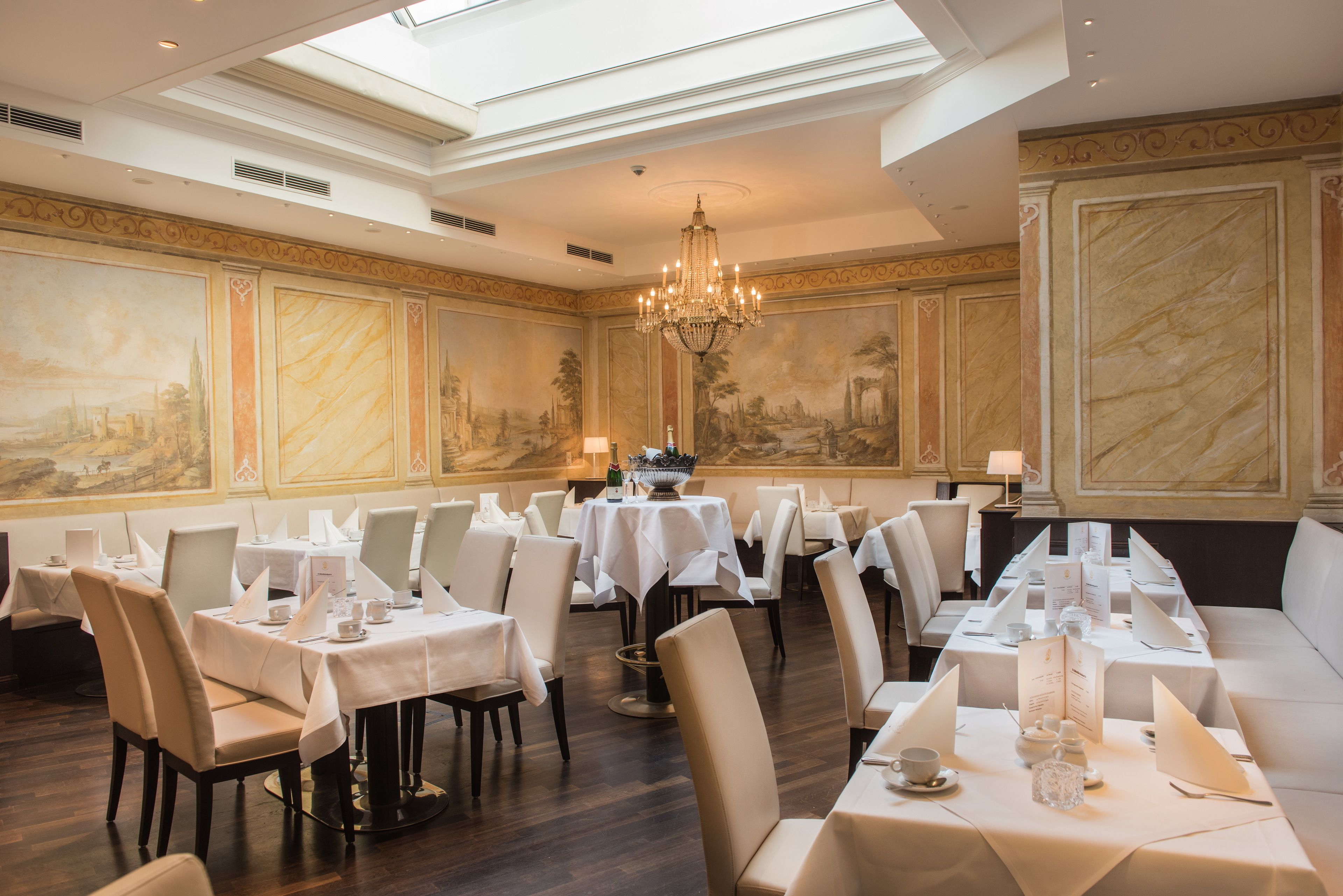 Une salle à manger élégante, avec des tables dressées pour le service, un lustre et des peintures murales de style classique.