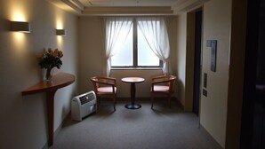 Habitació d'hotel amb una taula petita i dues cadires, un radiador portàtil, una finestra amb cortines lleugeres i un aire condicionat fixat a la paret.