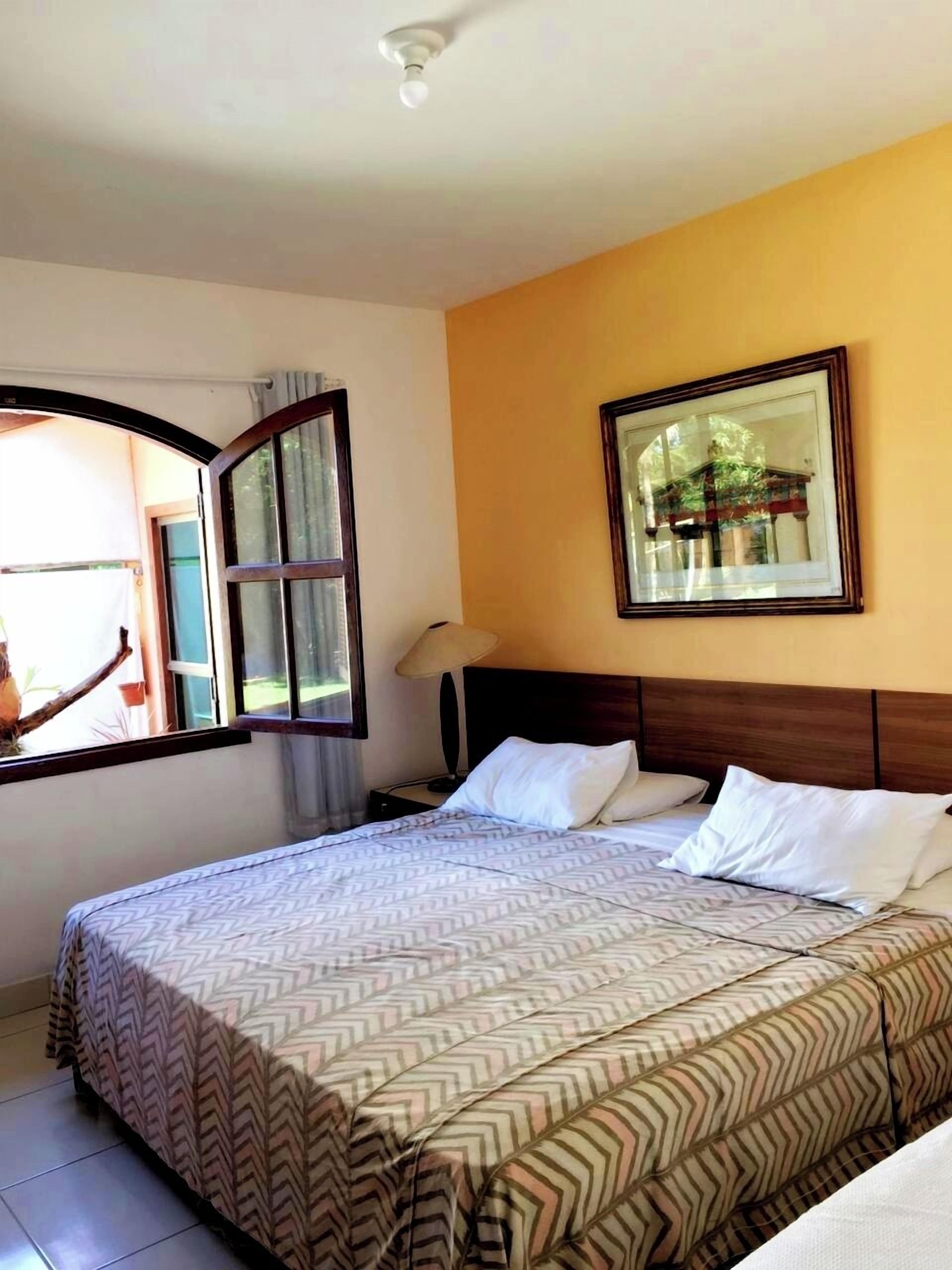 Une chambre d’hôtel avec un grand lit, une fenêtre avec des rideaux, une table de chevet, une lampe et un tableau encadré au mur.