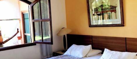 Une chambre d’hôtel avec un grand lit, une fenêtre avec des rideaux, une table de chevet, une lampe et un tableau encadré au mur.