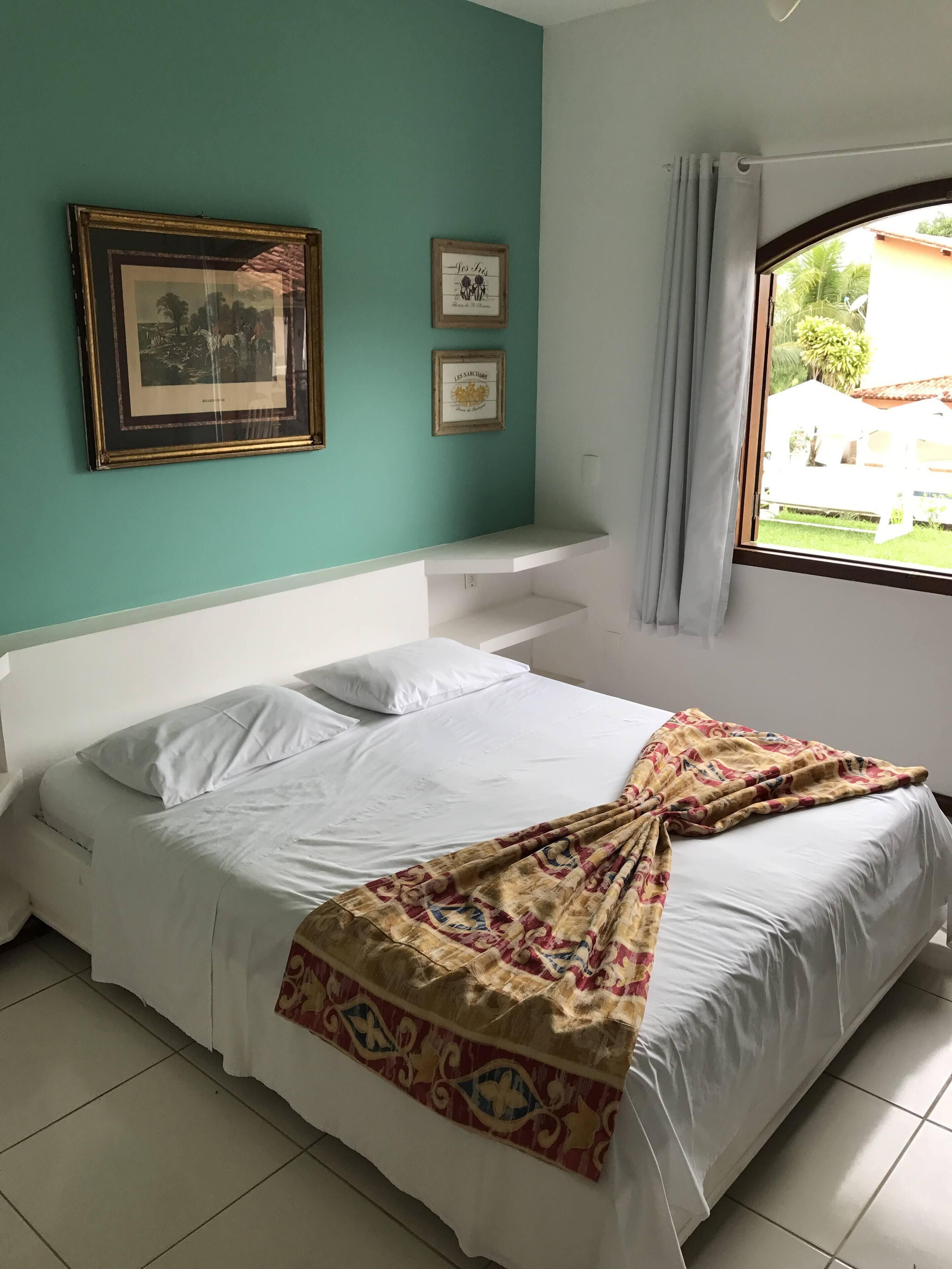 Een slaapkamer met een bed, een raam en twee foto’s in lijsten aan de muur.