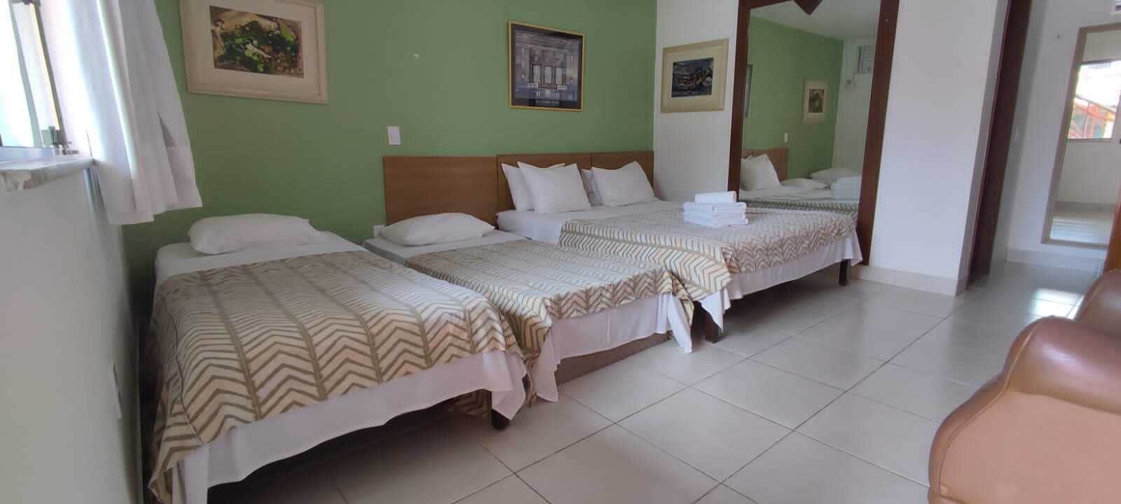 Hotelkamer met drie bedden, elk voorzien van een bedovertrek met een patroon en witte kussens. Er hangen foto's in lijsten aan de muur en er is een raam met gordijnen.