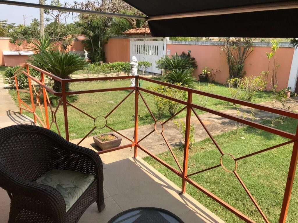 A balcony with a wicker chair, a small table, and a view of a garden with a fence and plants.