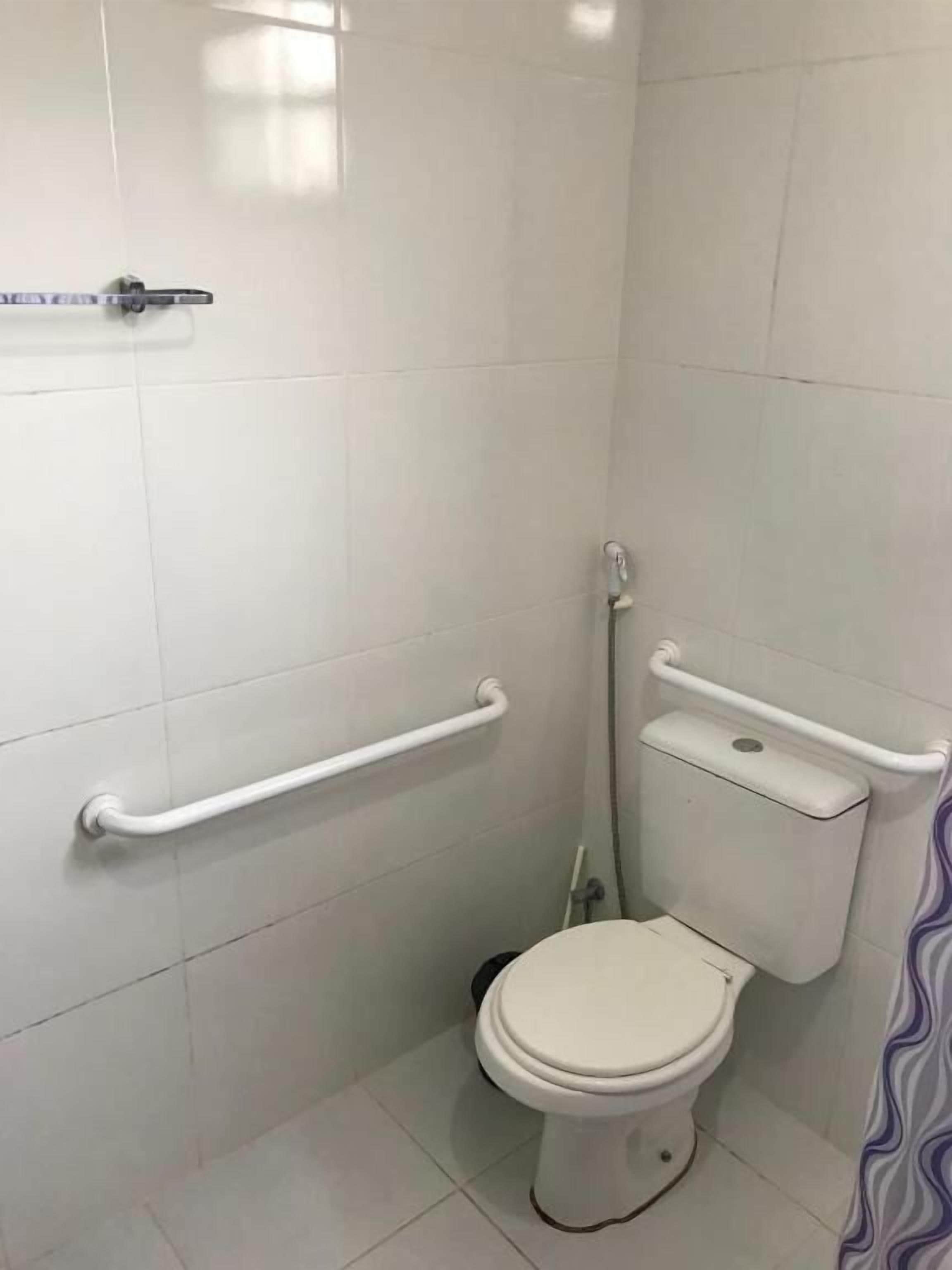 A small, white-tiled bathroom with a toilet, a handrail, and a grab bar.