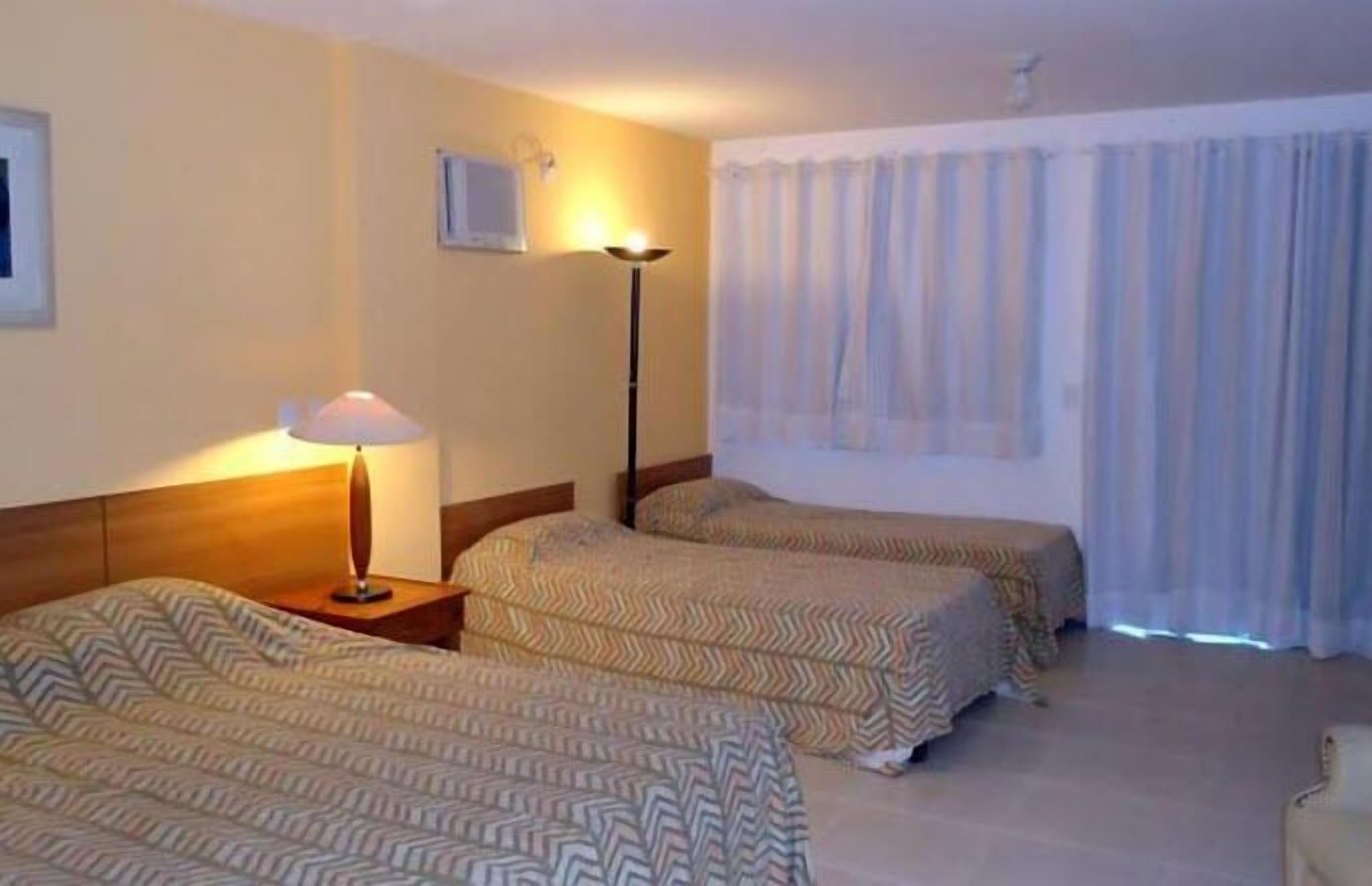 A hotel room with two single beds, a bedside table with a lamp, a window with curtains, and a wall-mounted TV.
