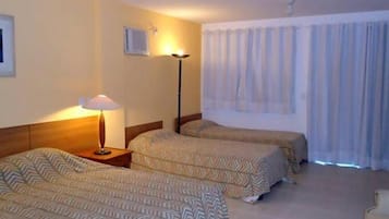 Quarto de hotel com duas camas de solteiro, mesa de cabeceira com abajur, janela com cortinas e televisão presa à parede.