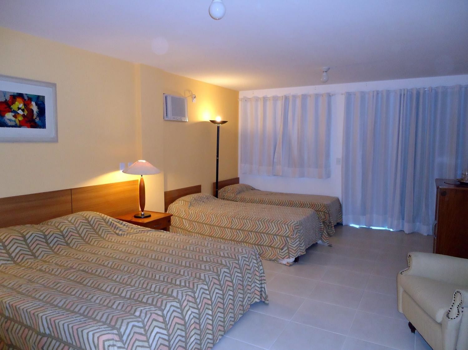 A hotel room with two beds, a nightstand, a lamp, a painting on the wall, and a window with curtains.