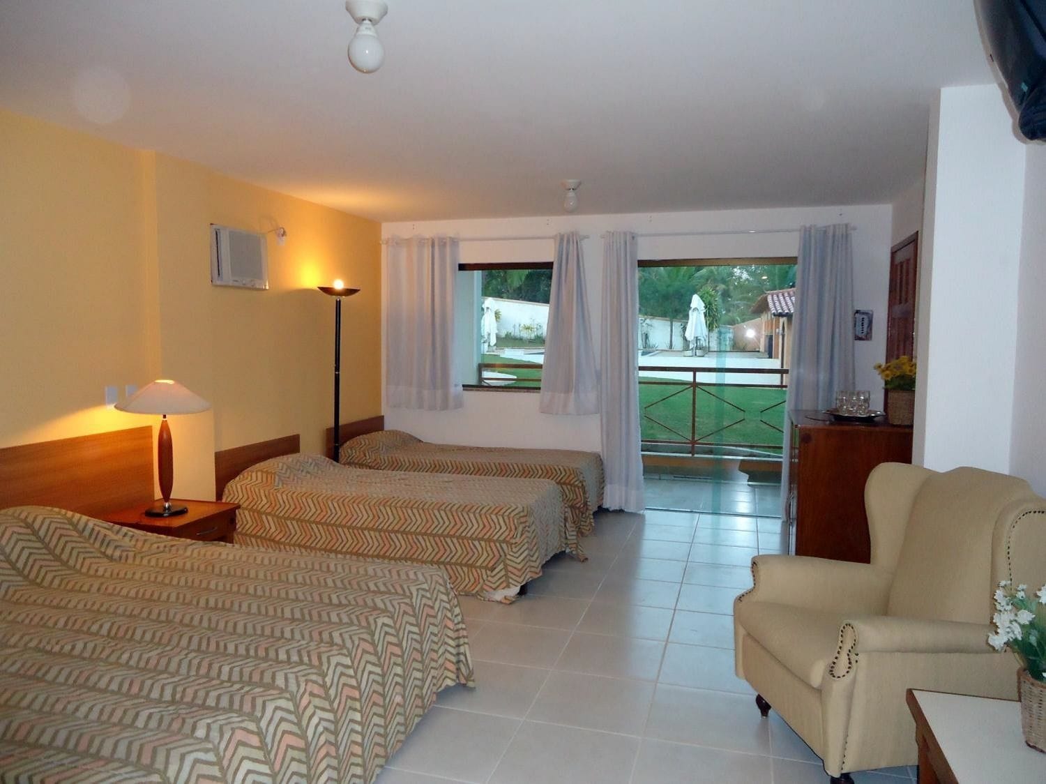 Habitación de hotel con dos camas, una silla, una lámpara y vistas al área exterior.