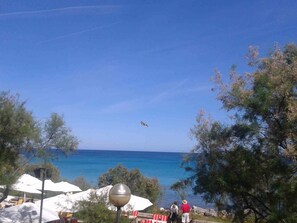 Vista costera con cielo despejado, una persona volando una cometa y algunas personas caminando por la orilla.