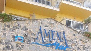 Eine Fassade aus Stein, mit dem Schild "AMENA" und einem großen Schaufenster, das den Himmel und einen Außenbereich mit Sitzgelegenheiten reflektiert.