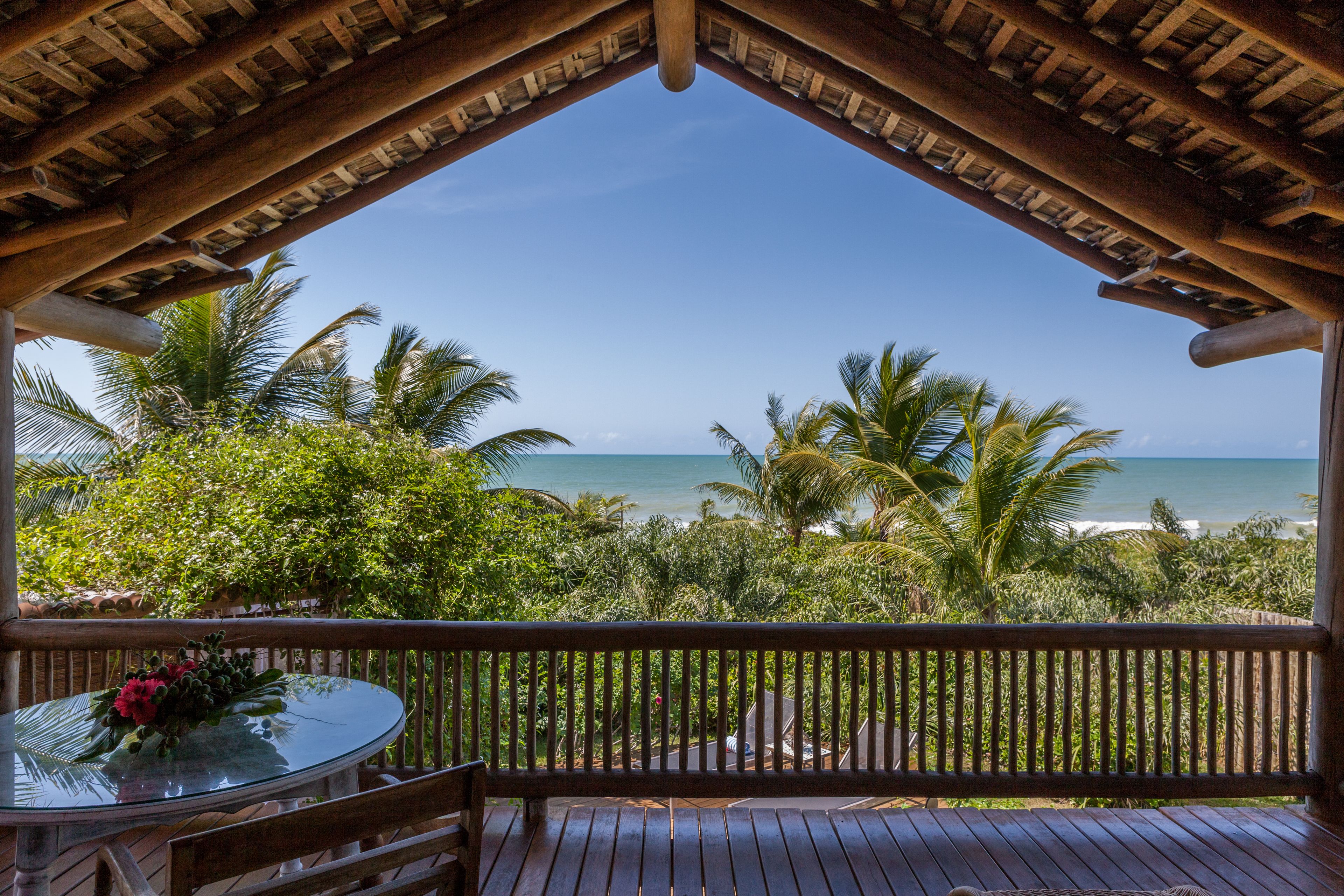 Una terraza de madera con una mesa de vidrio y un banco, con vista a una playa con palmeras y el océano.