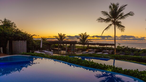 An infinity pool with a view of the sunset over the ocean, surrounded by lounge chairs and palm trees. - Pousada Estrela D'Água (Porto Seguro)