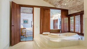A spacious bathroom with a large bathtub, wooden door frames, and a view of a bedroom with a bed and a chair. - Pousada Estrela D'Água (Porto Seguro)