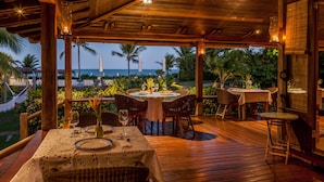 An outdoor dining area with wooden tables and chairs, overlooking a beach and palm trees. - Pousada Estrela D'Água (Porto Seguro)