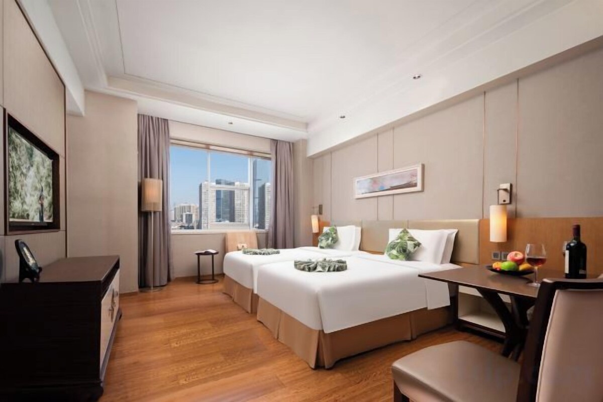Habitación de hotel con una cama grande, un escritorio, una silla, una ventana con vista a la ciudad y una mesa de comedor con una botella y fruta.
