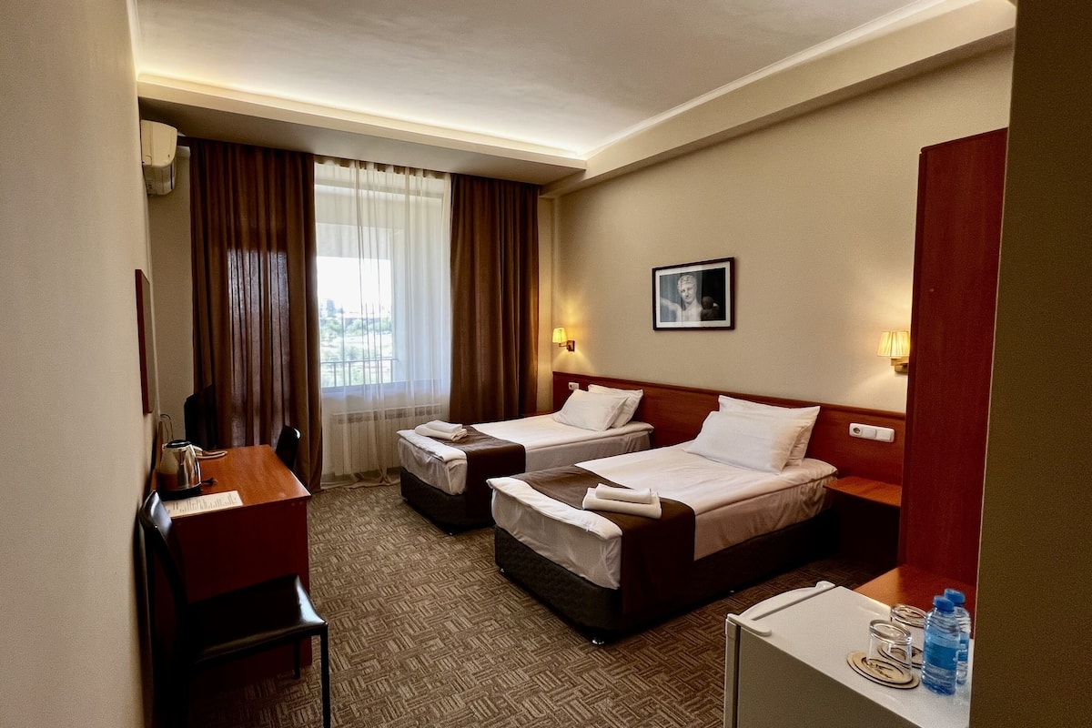 A hotel room with two beds, a desk, a chair, a small refrigerator, and a window with curtains.