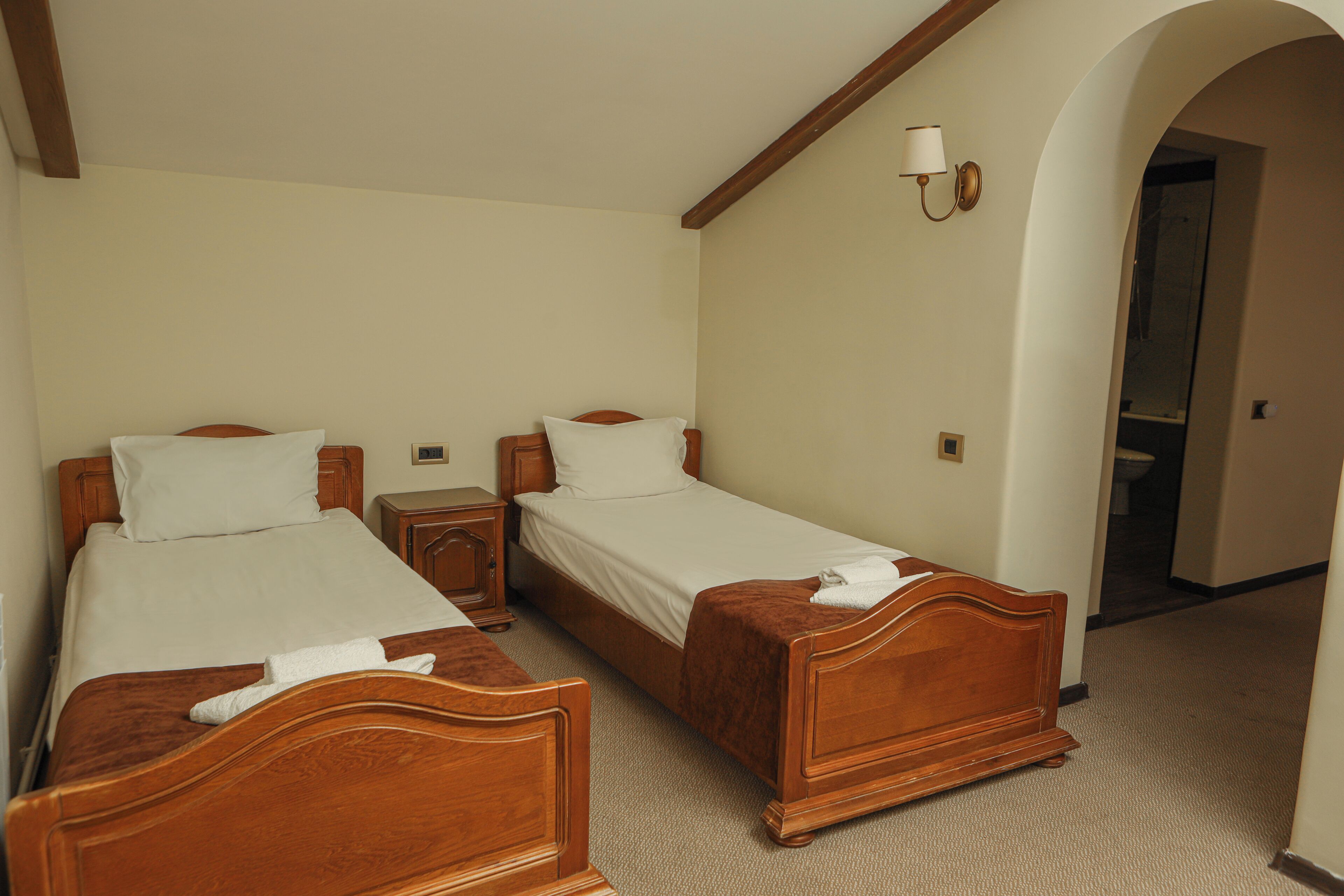 A hotel room with two wooden beds, a nightstand, a lamp, and a door leading to a bathroom.