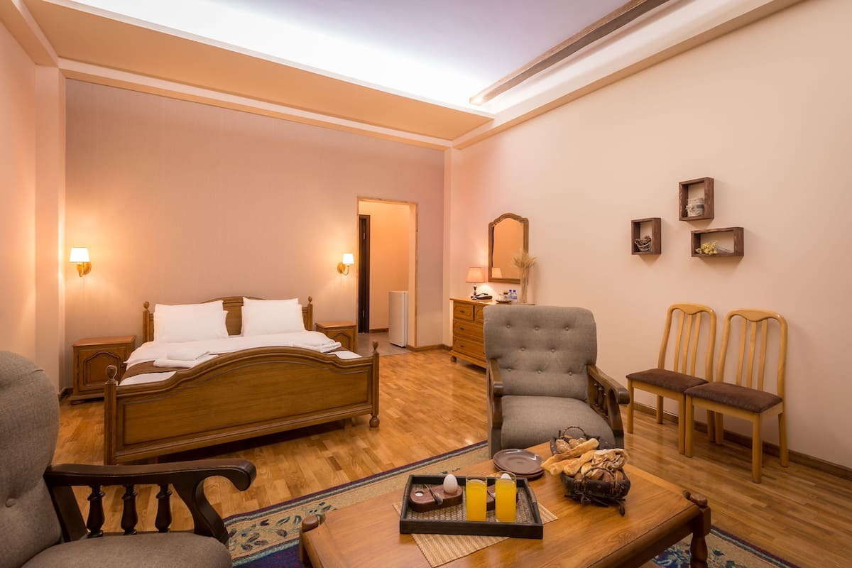 A hotel room with a wooden bed, a grey armchair, two wooden chairs, a small table with a tray of breakfast items, and a mirror on the wall.