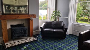 A room with a fireplace, a black leather sofa, and a plaid carpet. - Black Isle Holiday Apartments (Avoch)