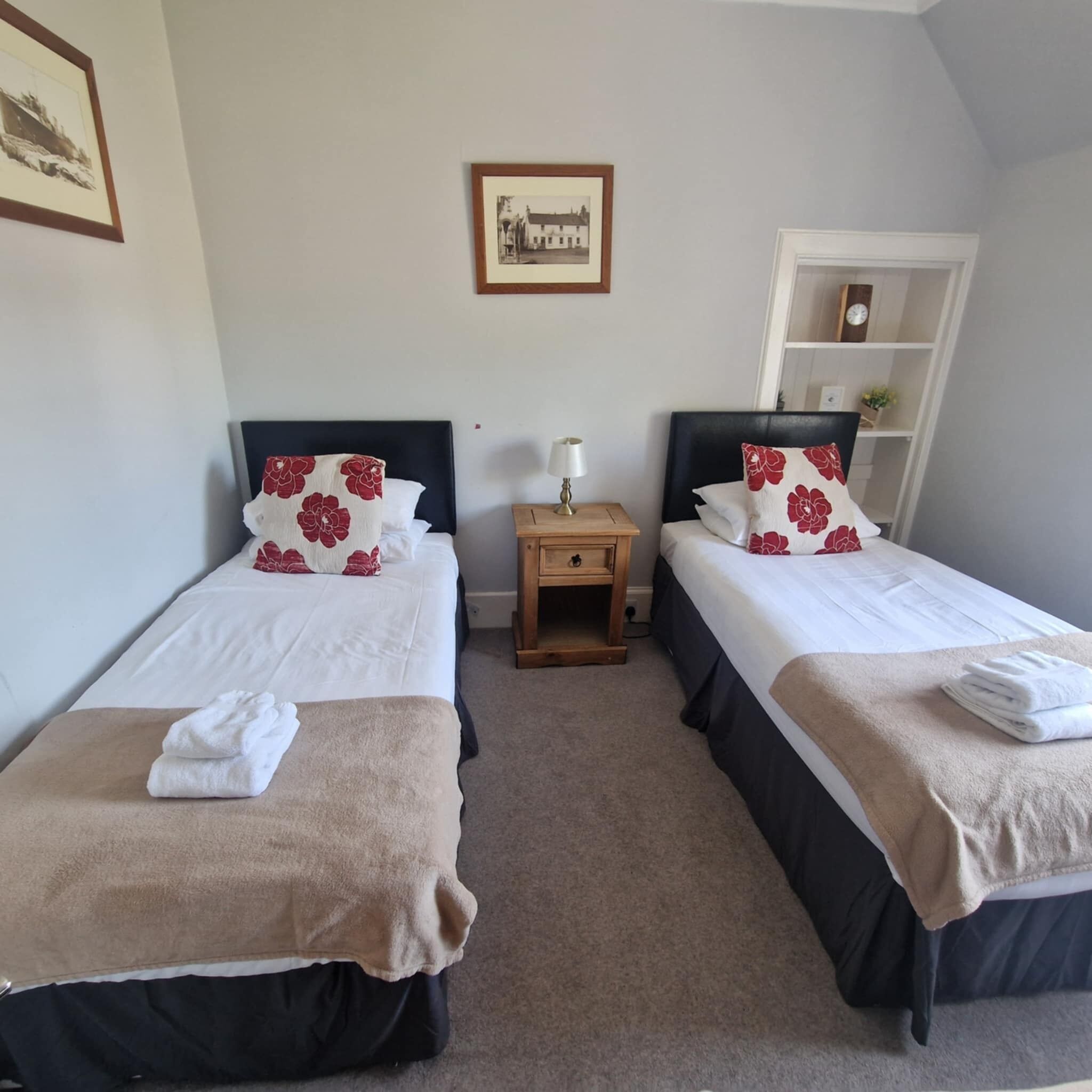 A hotel room with two single beds, each with a floral pillow and a beige blanket. There is a small wooden nightstand with a lamp between the beds.