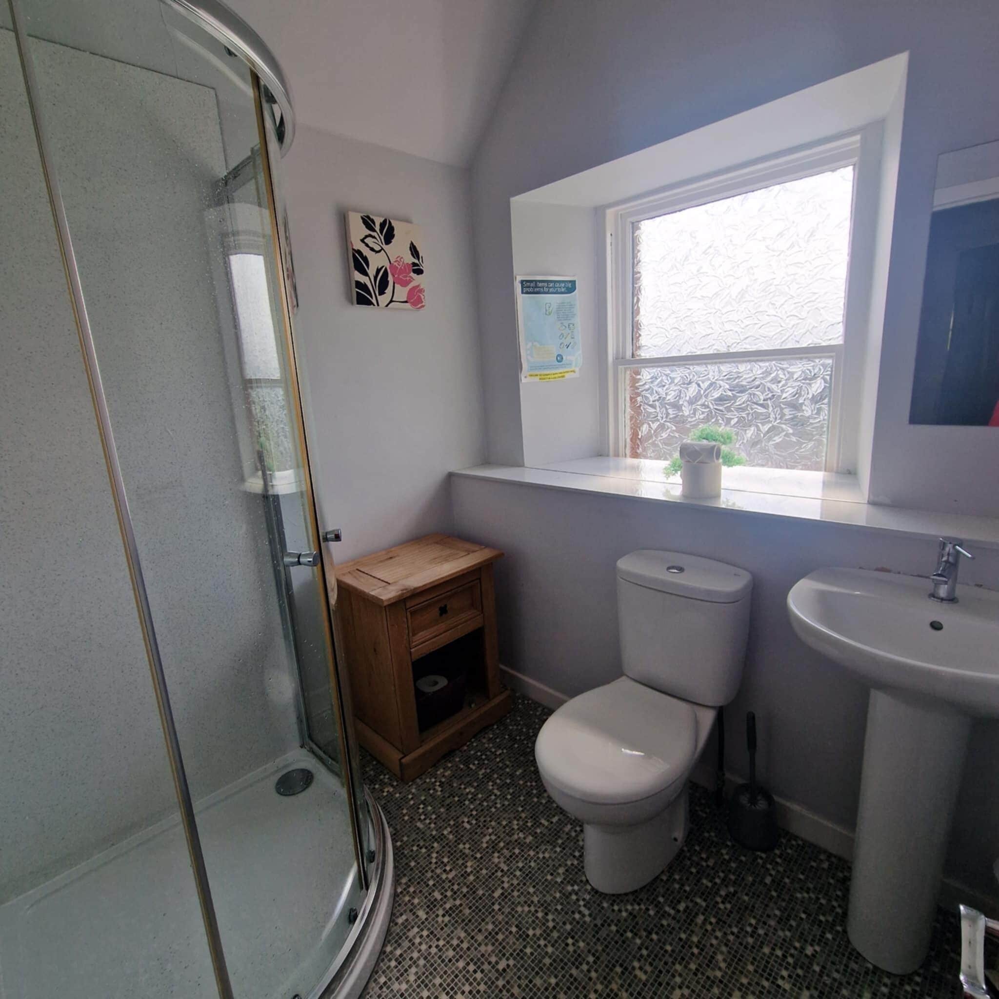 A bathroom with a glass shower enclosure, a toilet, a small wooden cabinet, a sink, and a window with frosted glass.