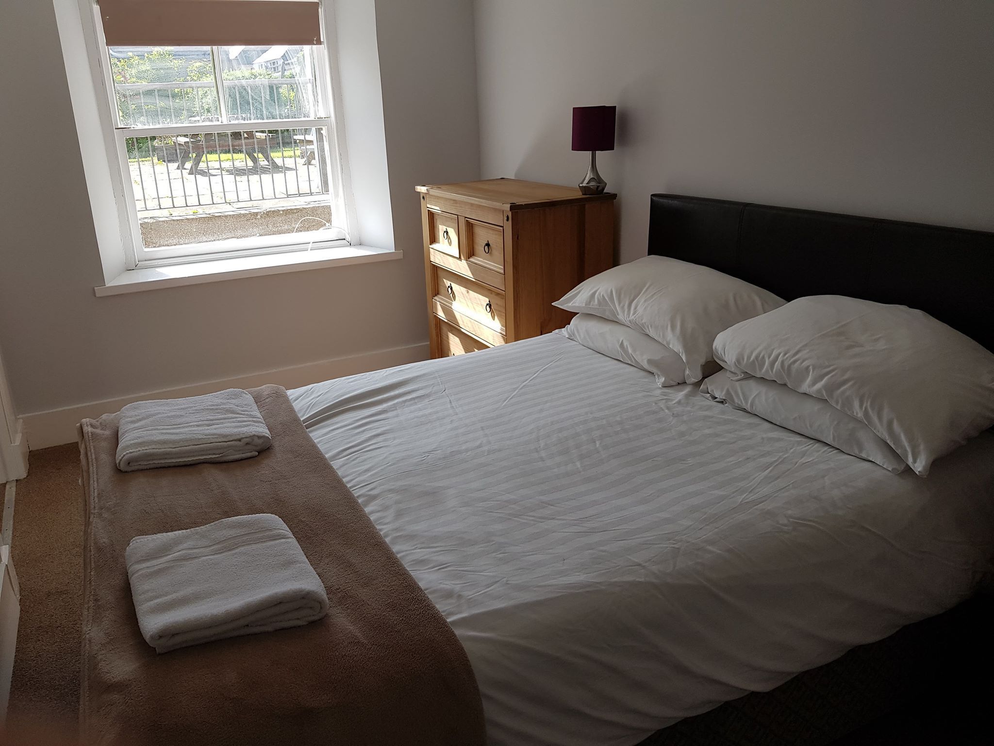 A bedroom with a bed, two pillows, a wooden nightstand, a lamp, and a window with a view of the outside.