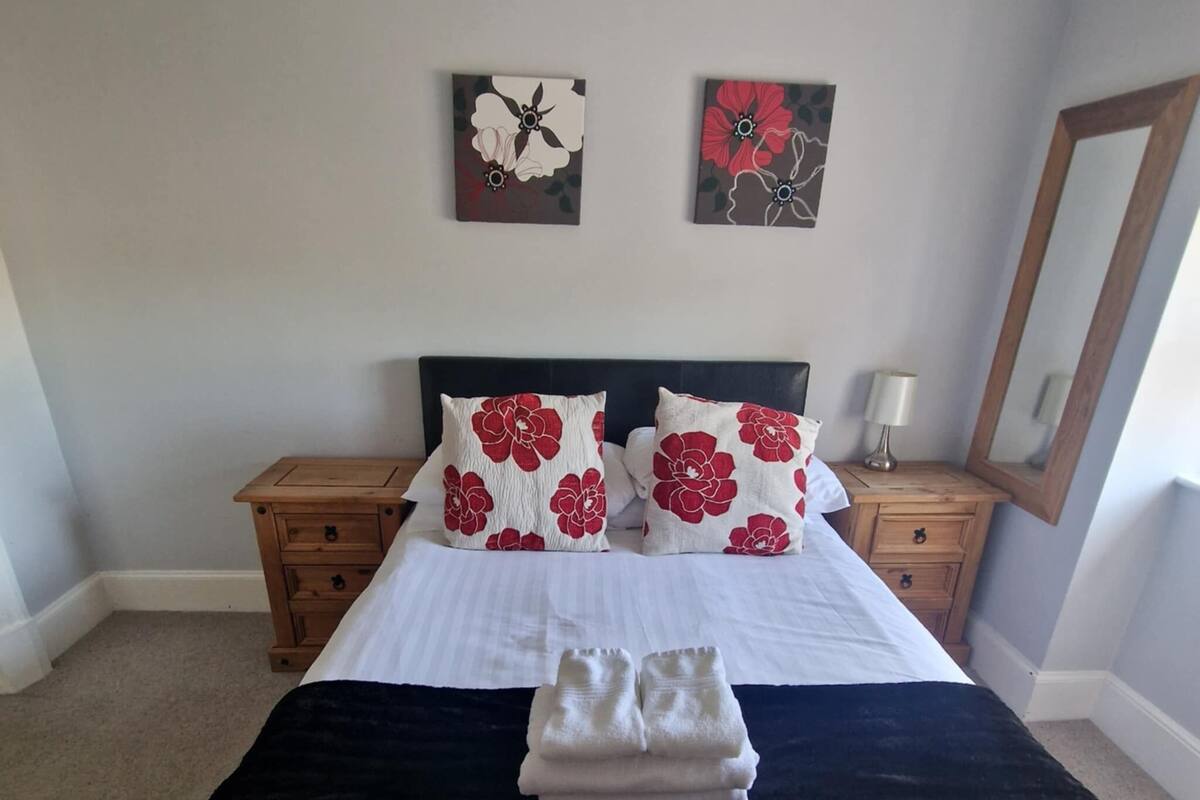 A neatly made bed with a black and white bedspread, two red floral pillows, and a folded white towel on the bed. There are two wooden nightstands with lamps, a large mirror on the wall, and two wall art pieces above the bed.
