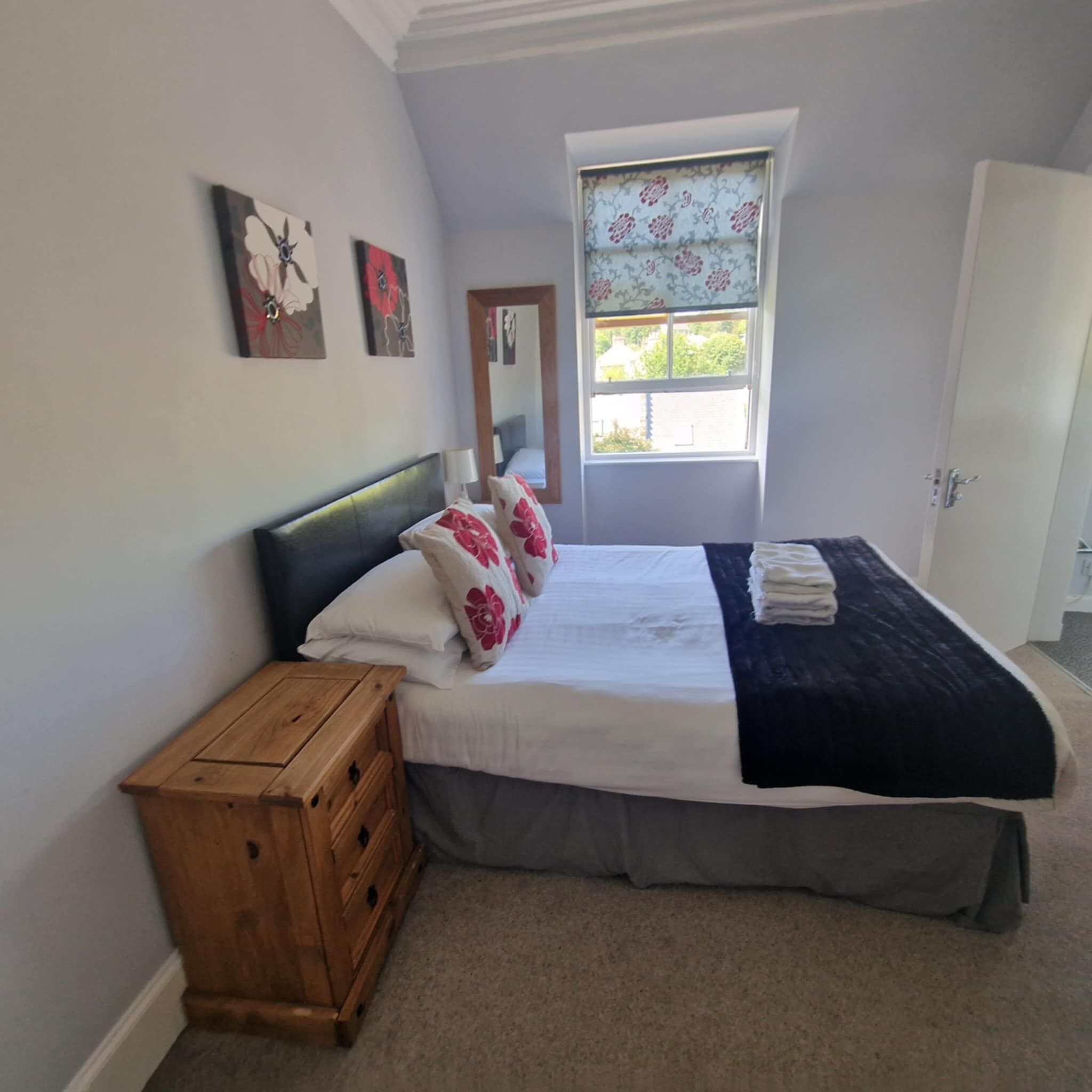Un dormitorio con una cama, una mesita de noche de madera, una ventana con cortinas florales y una puerta.