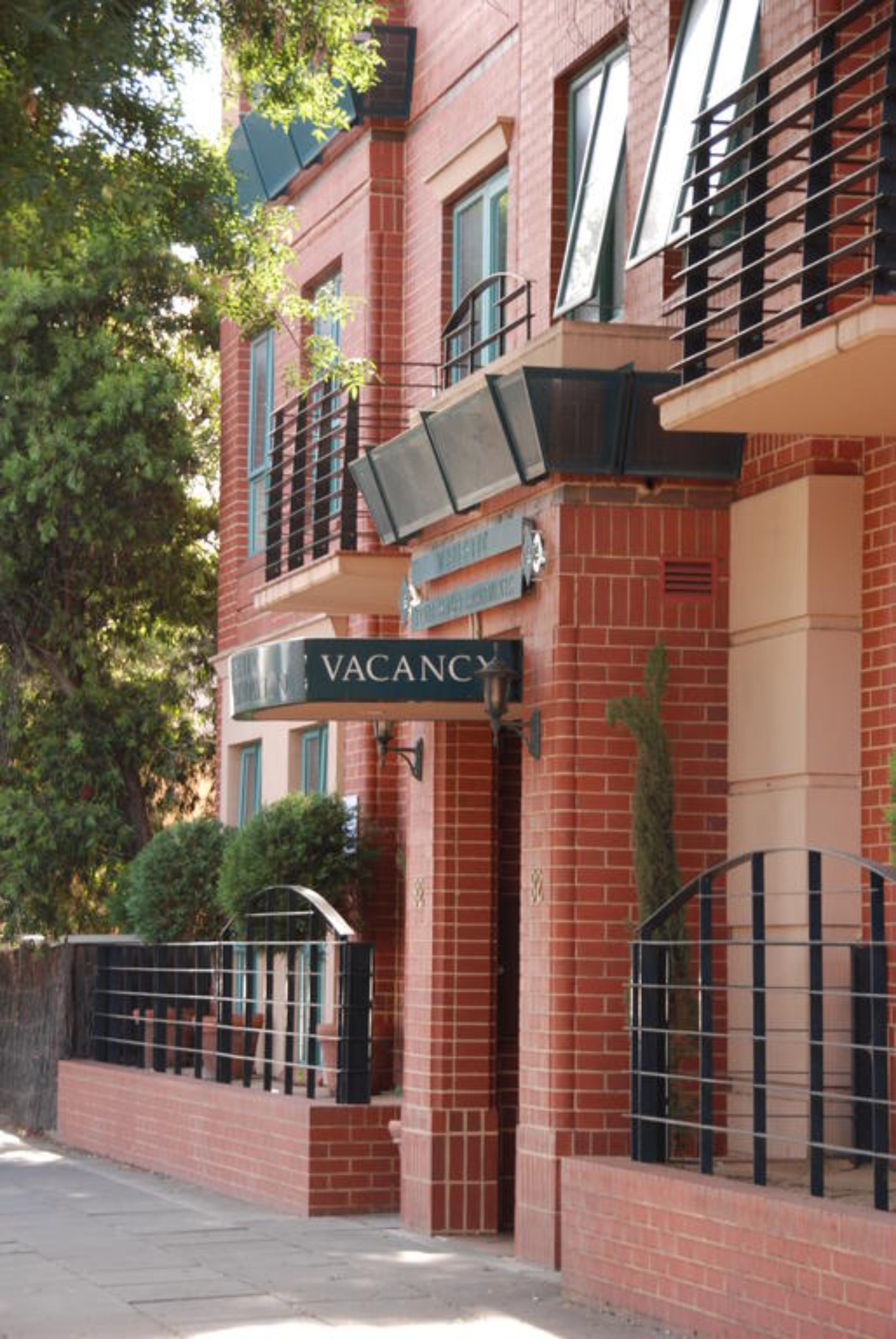 Un edificio de ladrillo rojo con un cartel de 'VACANCY', una entrada con rejas y un pequeño árbol en frente.