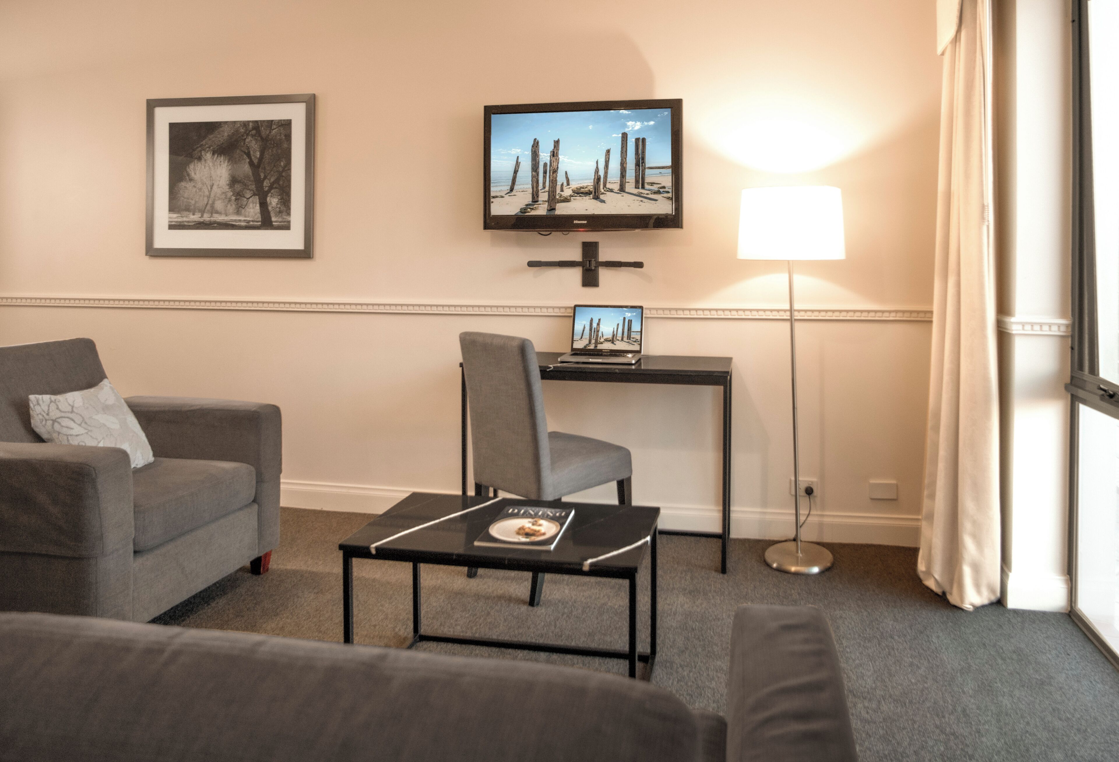 A hotel room with a sofa, a chair, a small table with a magazine, a TV on a stand, and a floor lamp.
