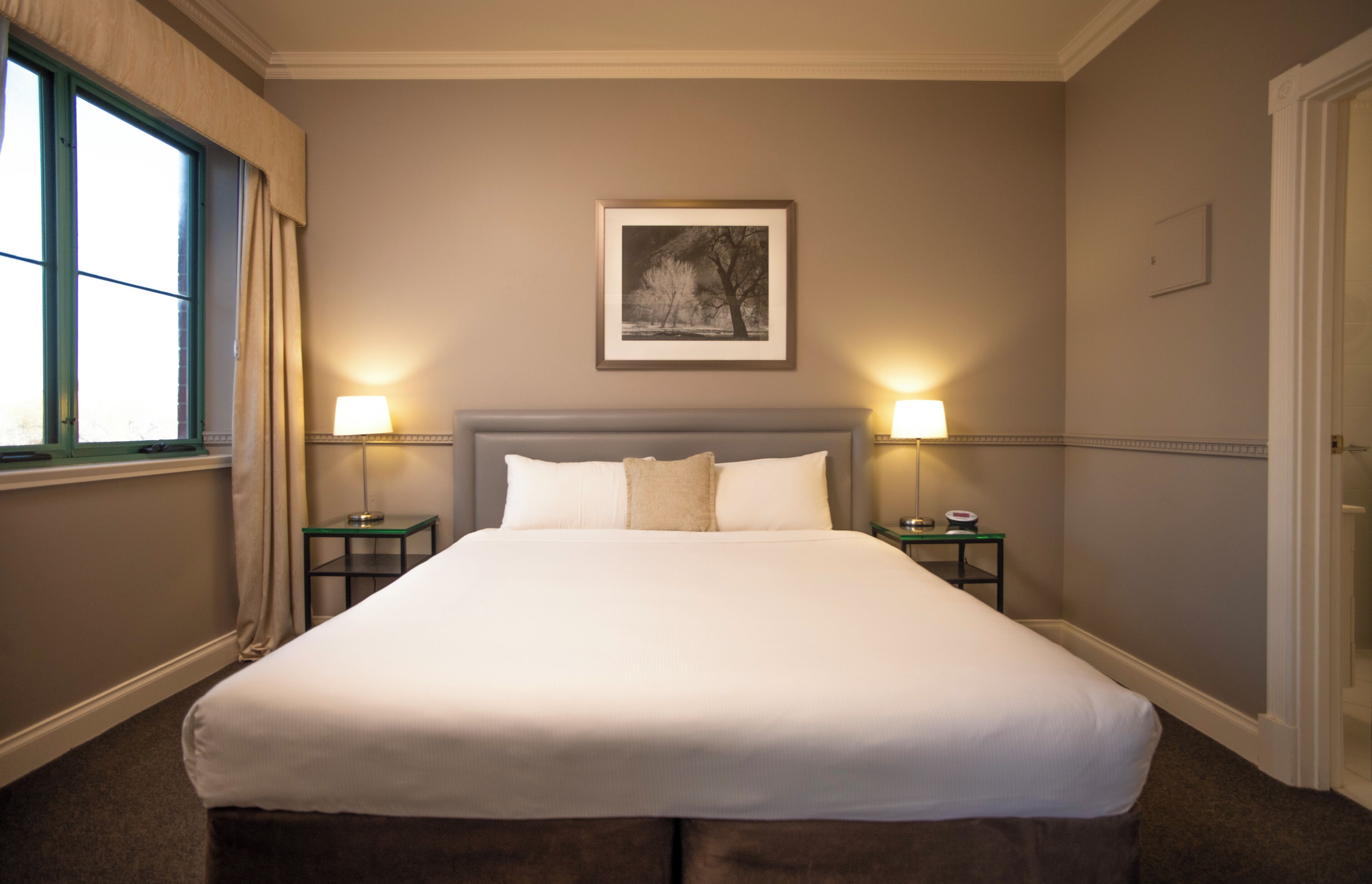 Habitación de hotel con una cama grande, dos mesitas de noche, un cuadro en la pared y una ventana con cortinas.