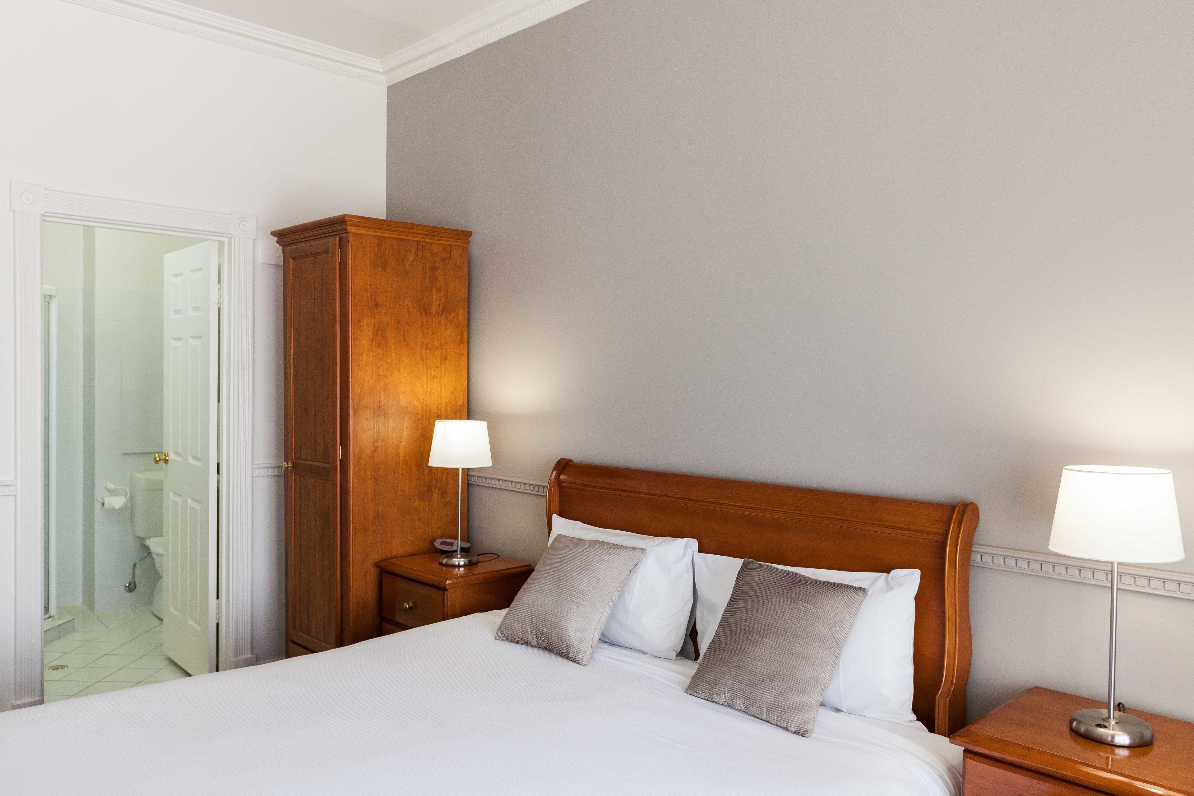 Une chambre d’hôtel avec une tête de lit en bois, un lit avec du linge de maison blanc, deux tables de chevet avec des lampes et une armoire.