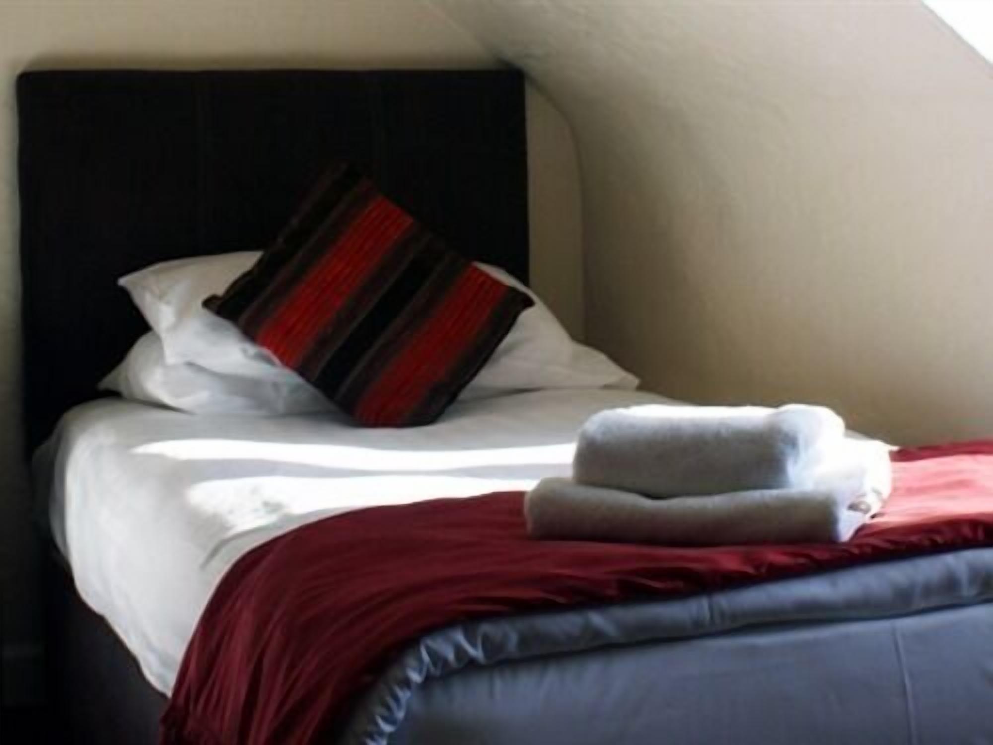 A neatly made bed with white linens, a red and black striped pillow, and a folded white towel.