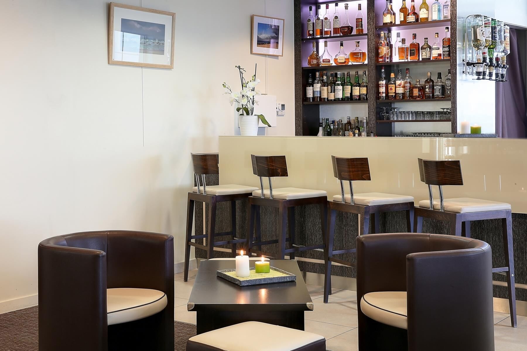 Un espace bar avec de nombreux tabourets et une table décorée de bougies. Des tableaux encadrés ornent le mur, et une étagère expose diverses bouteilles.