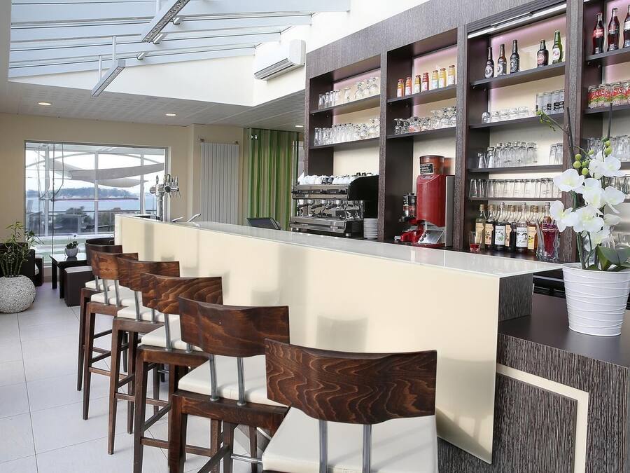 A modern bar area with a long counter, multiple stools, and shelves stocked with bottles and glasses.