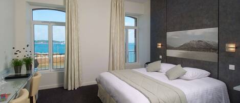 A hotel room with a large bed, a desk with a chair, a view of the sea, and a mountain landscape on the wall.