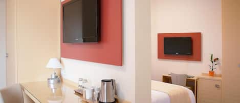 Ett hotellrum med en säng, ett skrivbord med en lampa, en kaffebryggare och två platt-TV-apparater som är monterade på väggen.