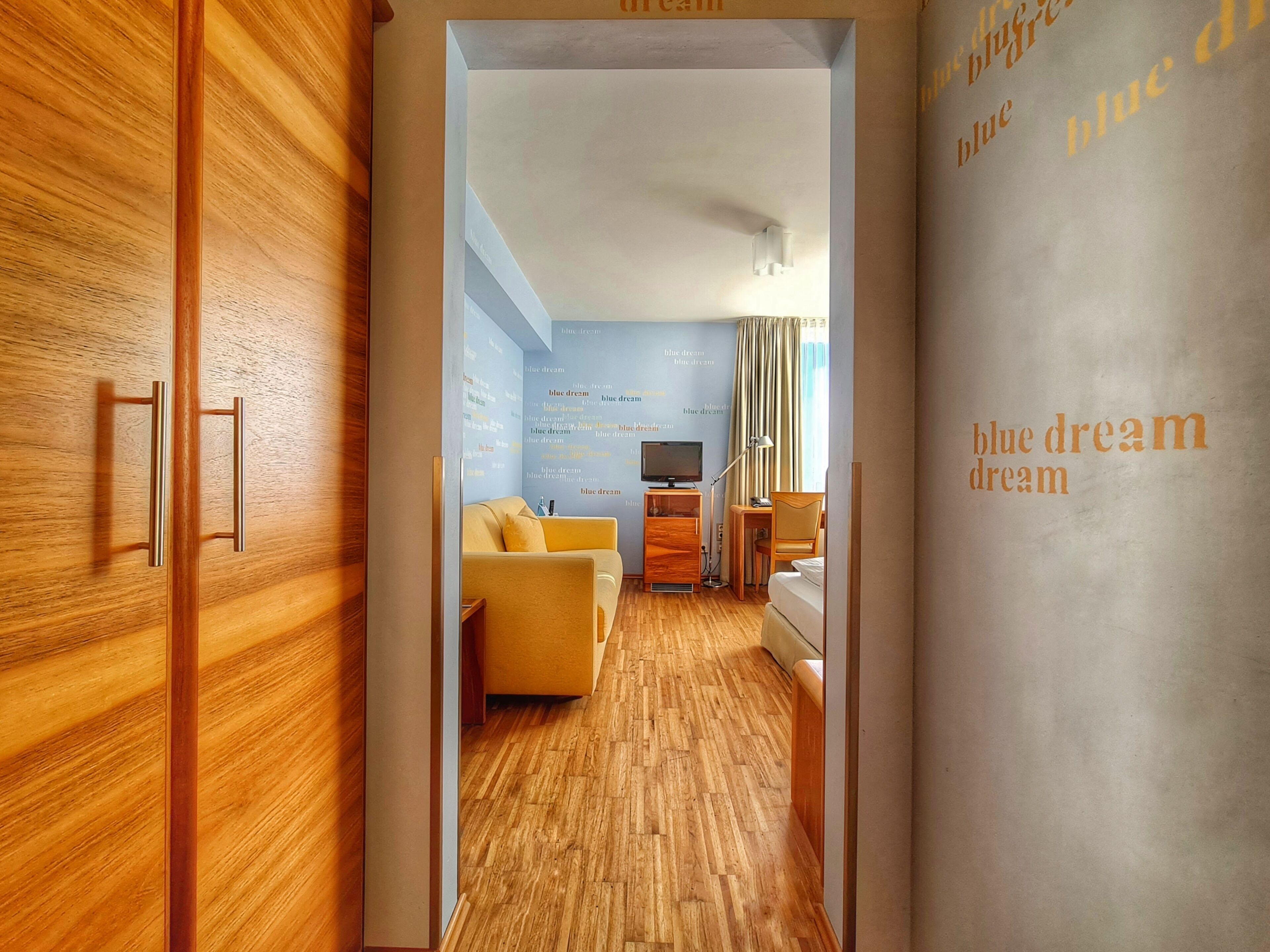 Habitación de hotel con suelo de madera, un sofá amarillo, televisor y cama.