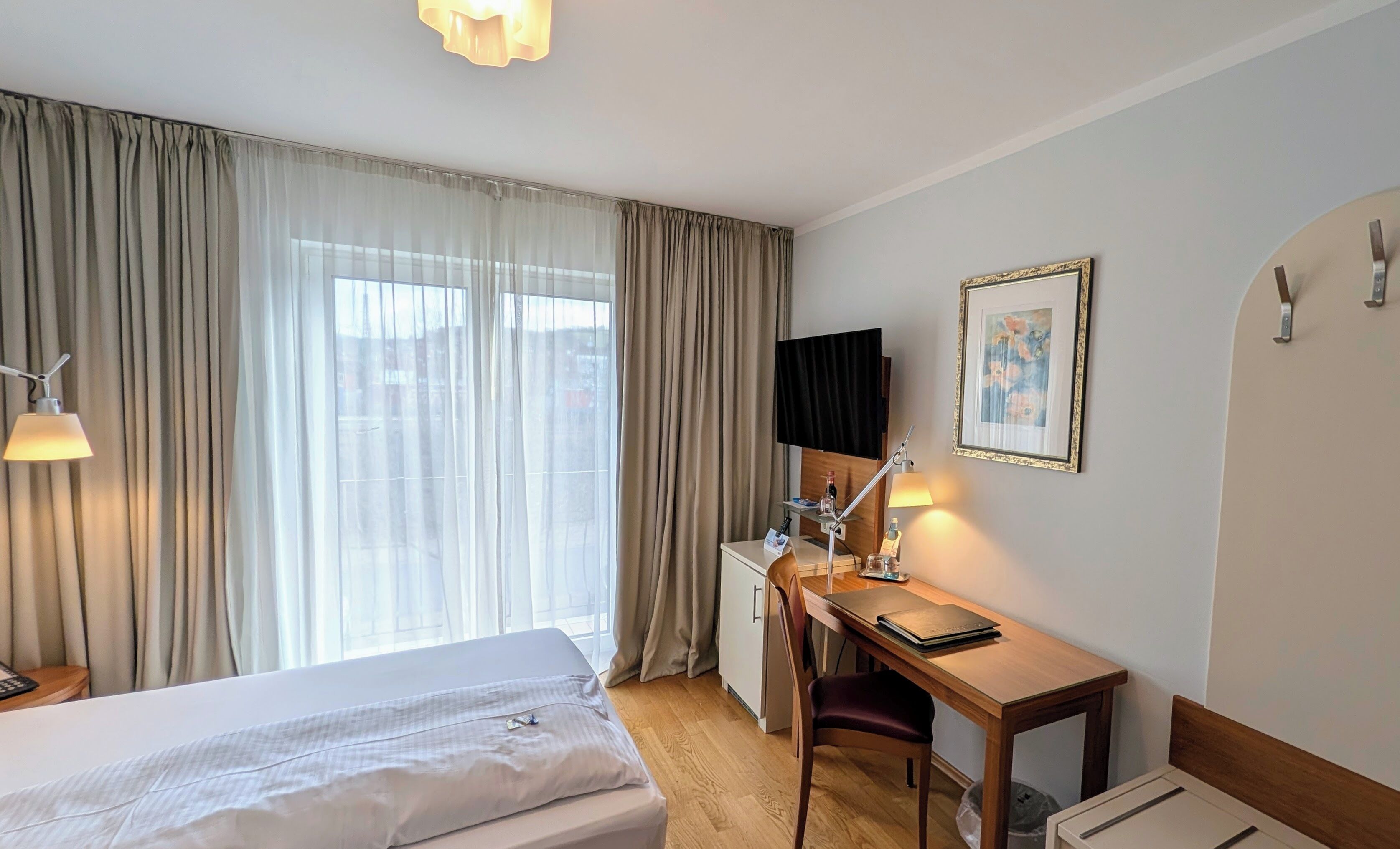 Ein Hotelzimmer mit Bett, Schreibtisch, Sessel, Fernseher und einem gerahmten Bild an der Wand.