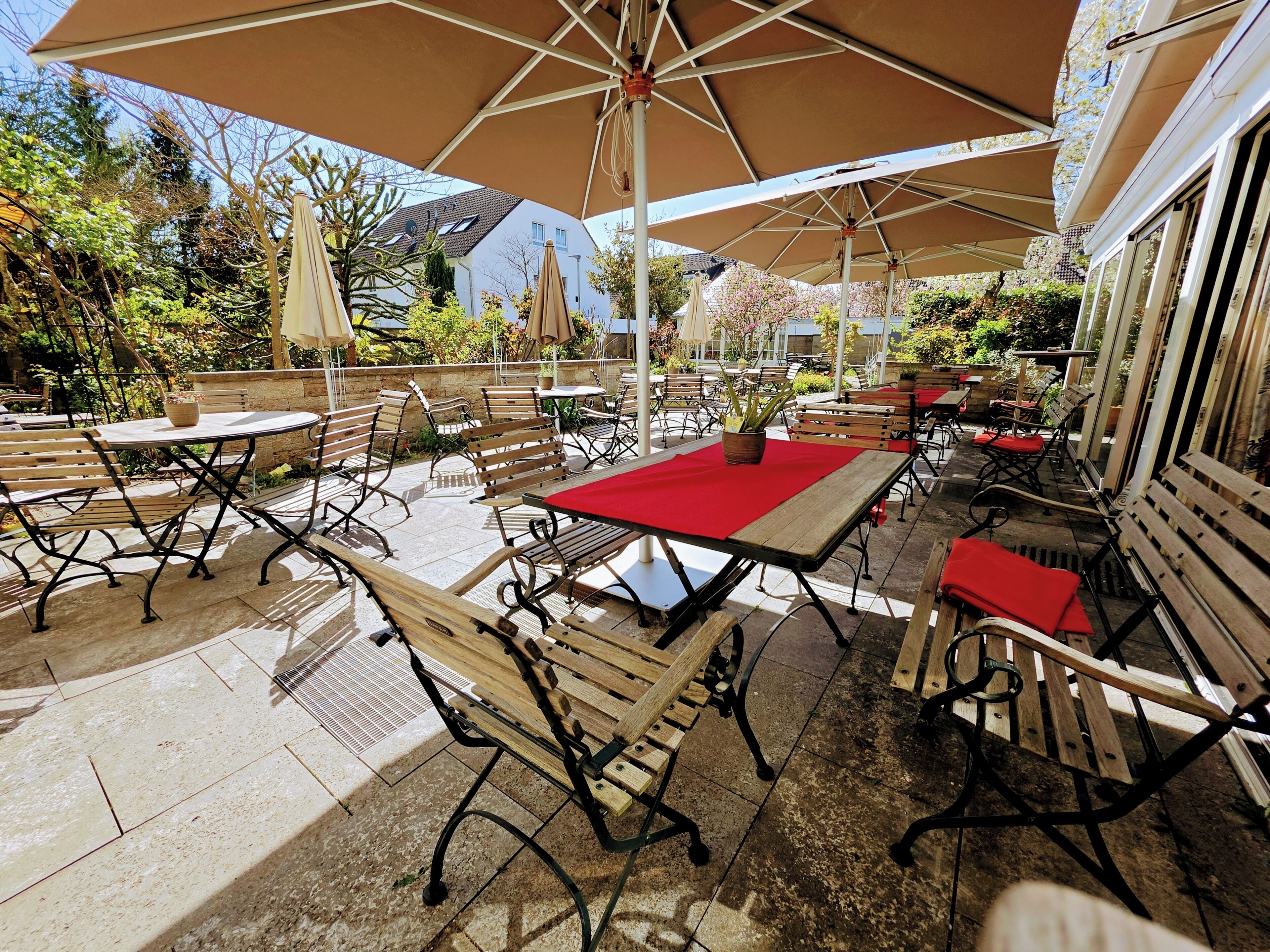 Un área de patio al aire libre con varias mesas y bancos, algunos con cojines rojos, bajo grandes sombrillas.