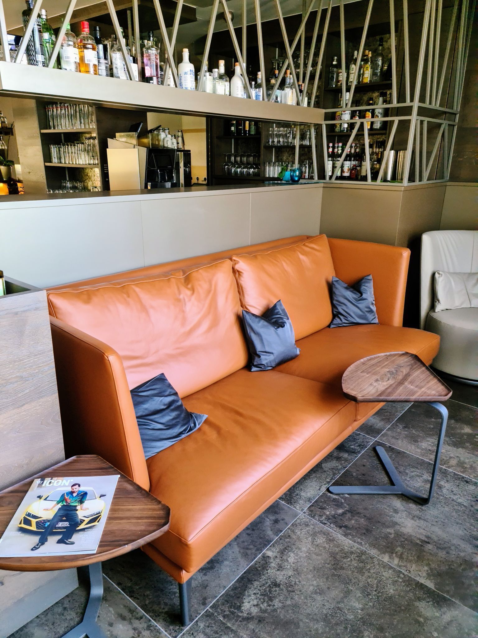 A modern bar area with a leather sofa, a small wooden table with a magazine, and a bar counter with various bottles and glasses.