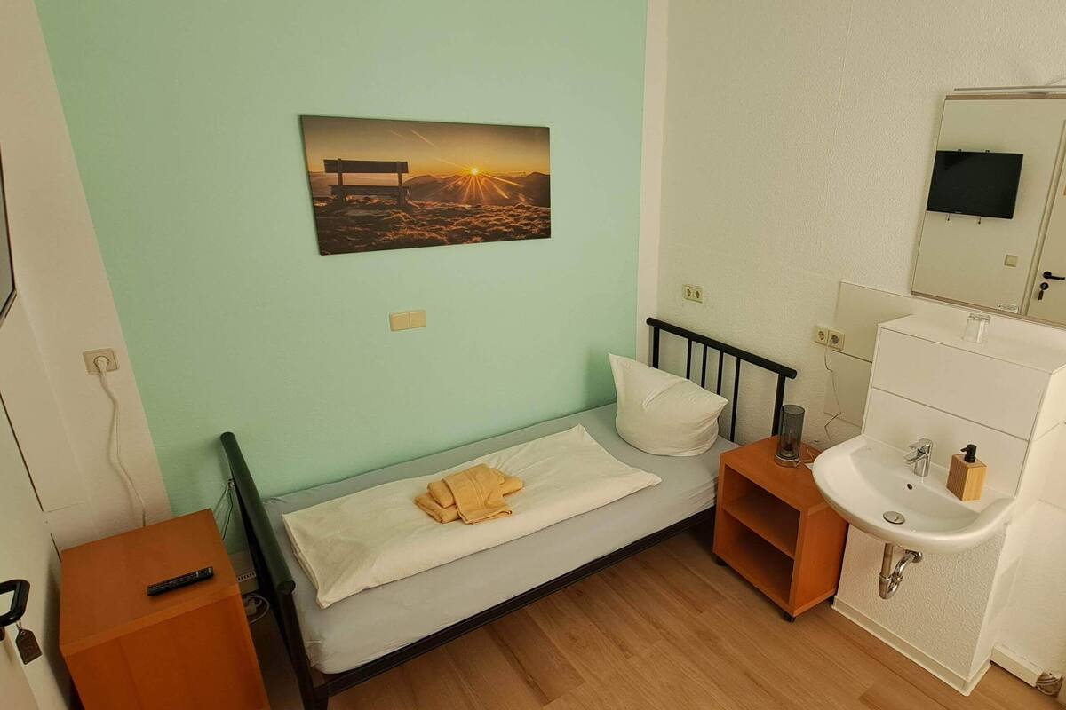 A small, neatly organized bedroom with a single bed, a bedside table, a nightstand, a wall-mounted TV, a bedside lamp, and a bathroom with a sink and mirror.
