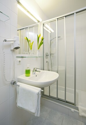 Un baño moderno con cabina de ducha de vidrio, un lavabo blanco con repisa de vidrio encima y un perchero con una toalla blanca.