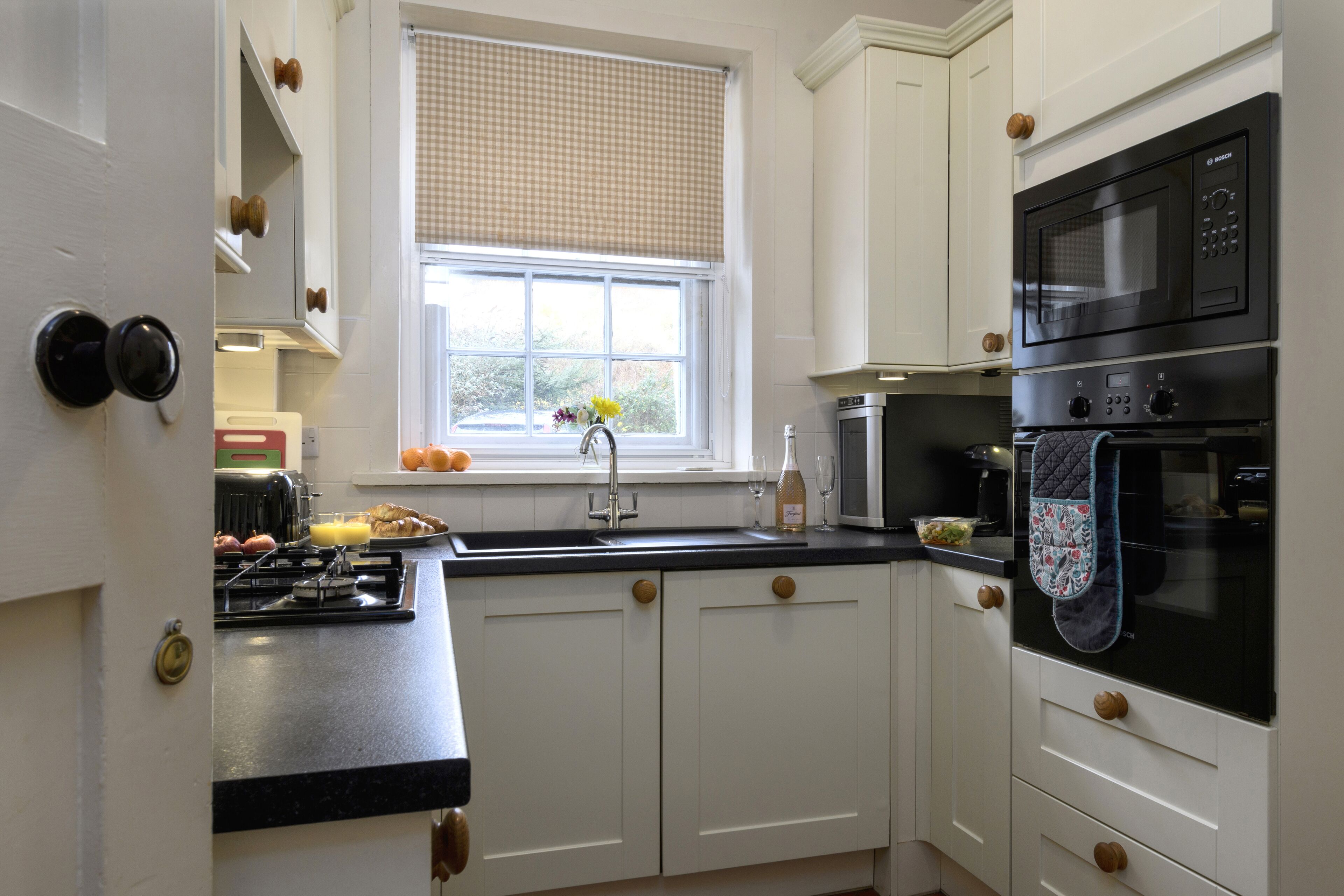 A kitchen with white cabinets, a black countertop, a window with a checkered blind, and a microwave oven.