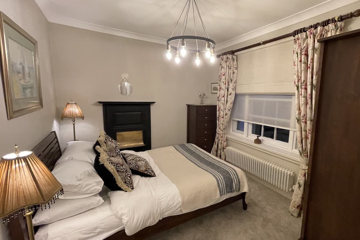 Um quarto com cama, mesa de cabeceira, cômoda, radiador e janela com cortinas.