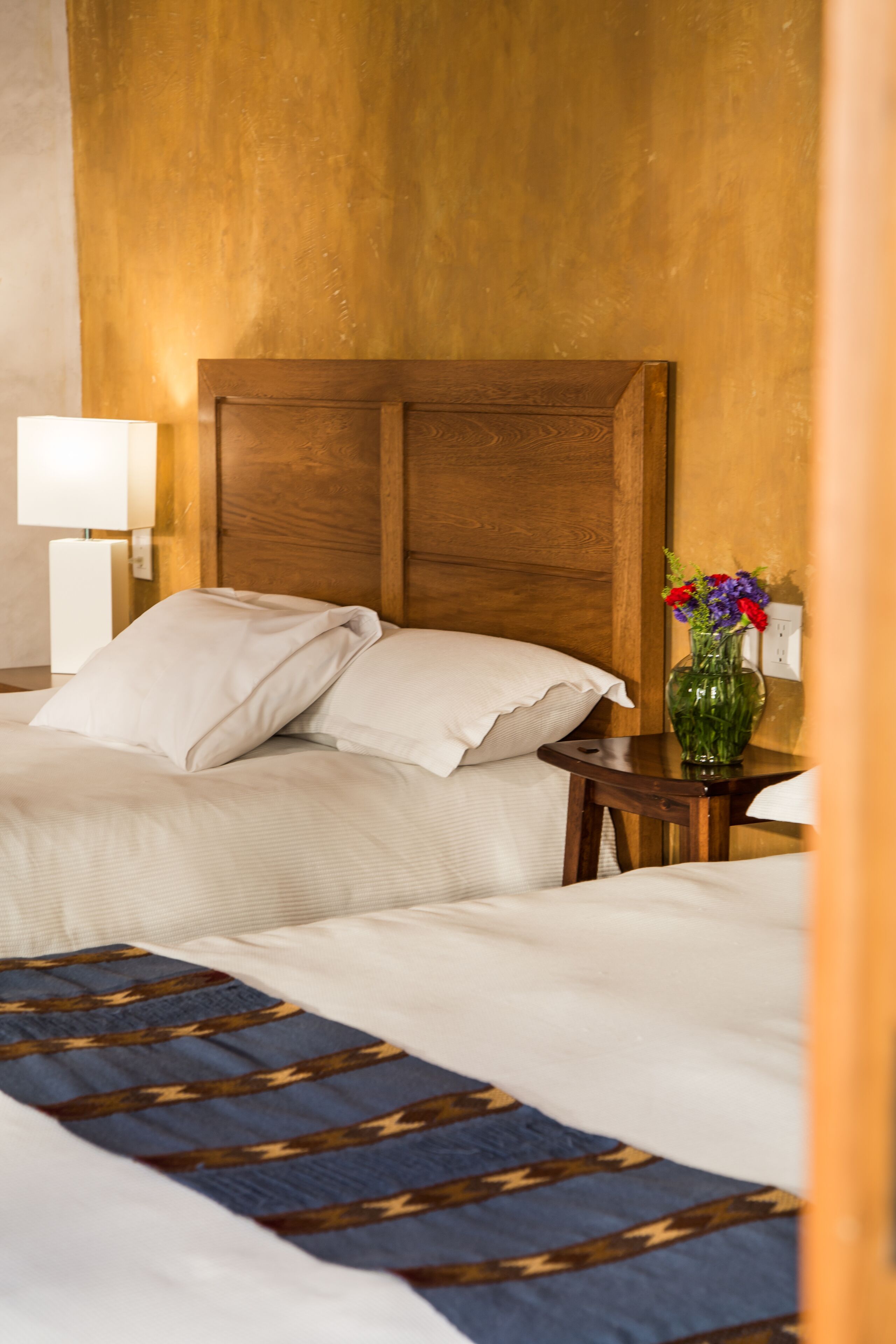 A hotel room with a wooden headboard, two beds, a nightstand with a lamp, and a vase with flowers.