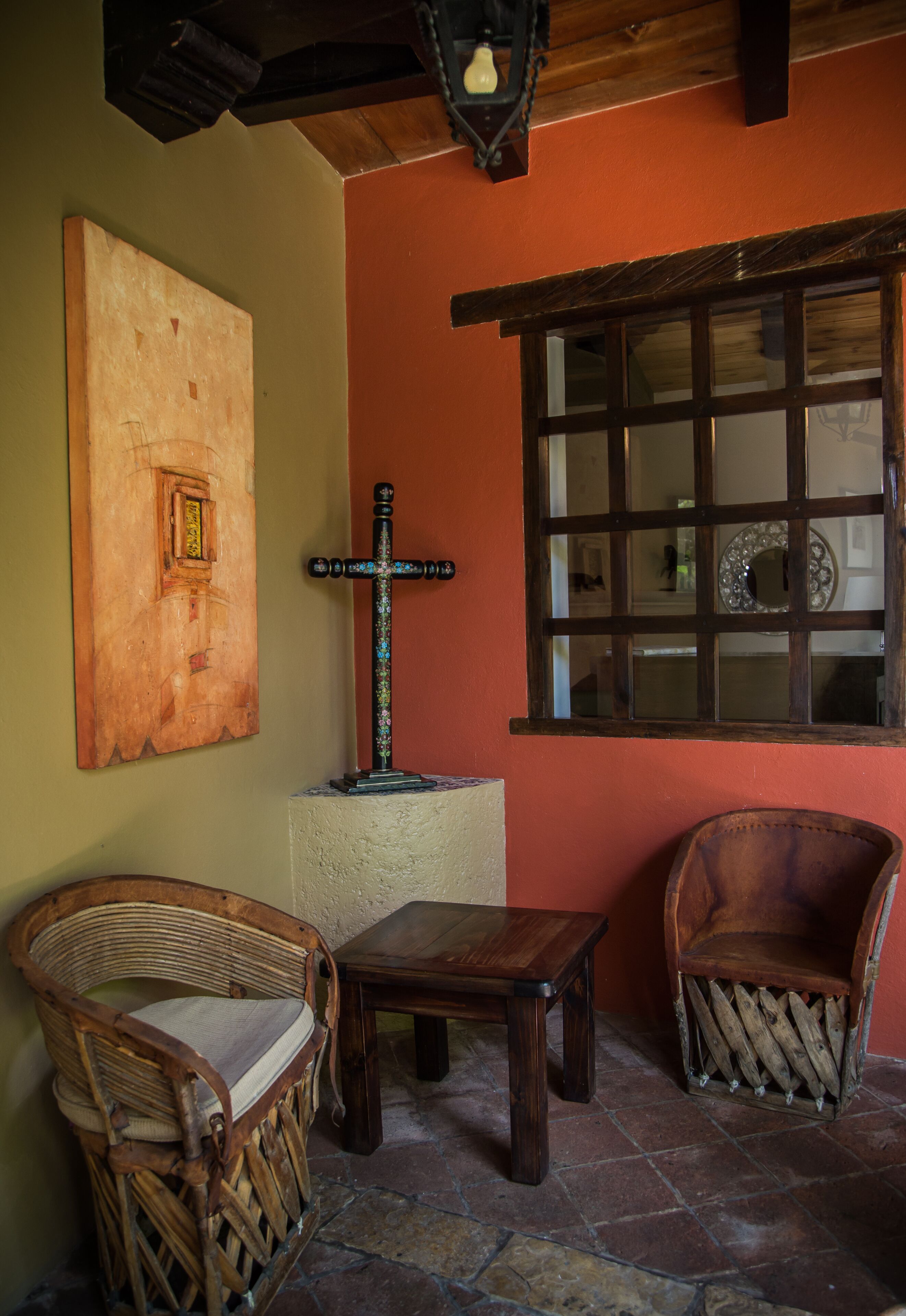 Una habitación con una mesa de madera, dos sillas de mimbre, una cruz de madera y una ventana con un espejo decorativo.
